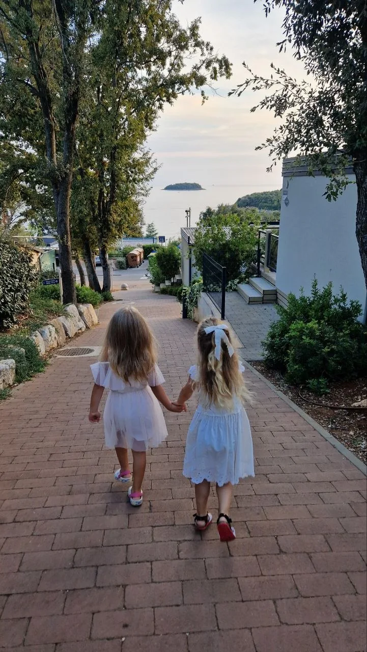 Two little girls in white dresses walking hand in hand down a paved path towards the sea, shaded by trees
