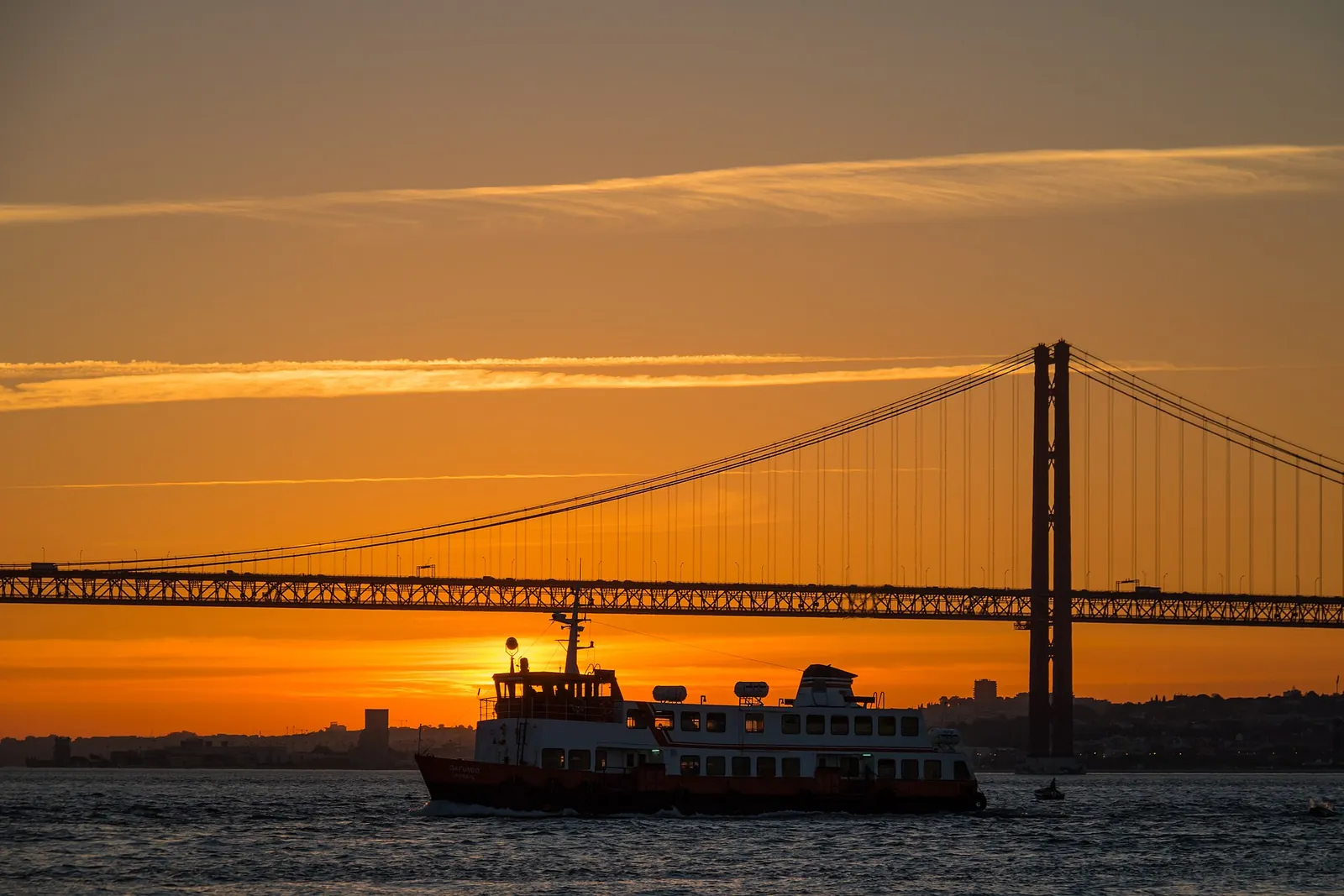 Ferry passing the 25 de Abril Bridge at sunset