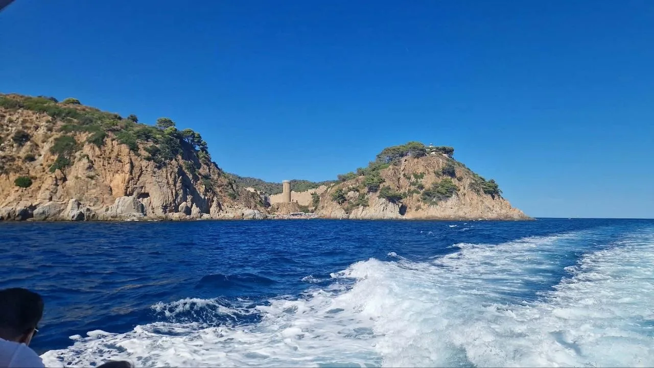 The rocky headland of Tossa de Mar and its old town walls, seen from a boat with its white wake in the blue sea
