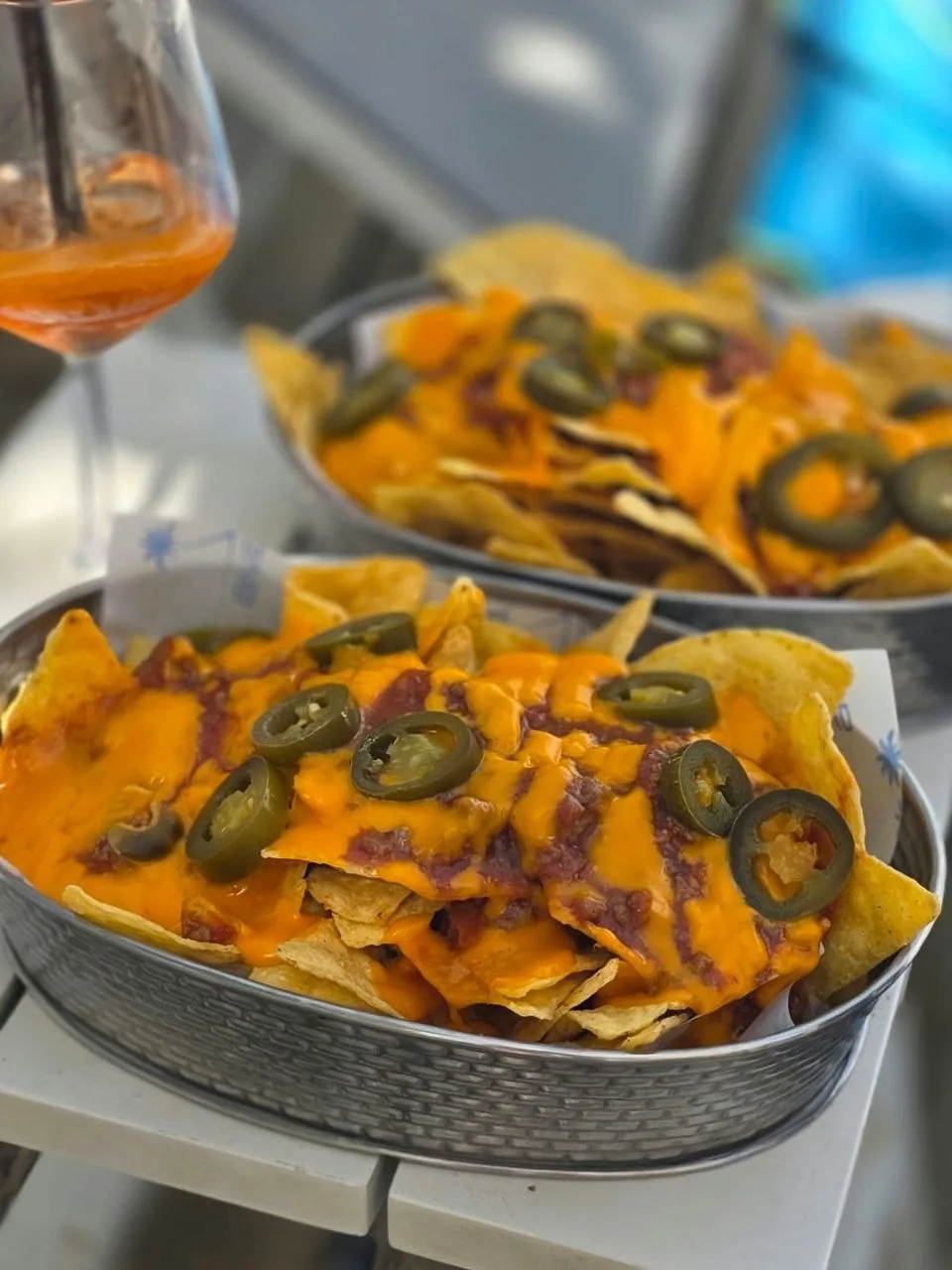 Plates of nachos with melted cheese and jalapeños and a glass of spritz on a bar table