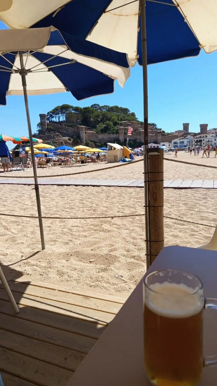 Tossa de Mar's castle and walls behind the beach, seen from a beach bar under blue and white umbrellas