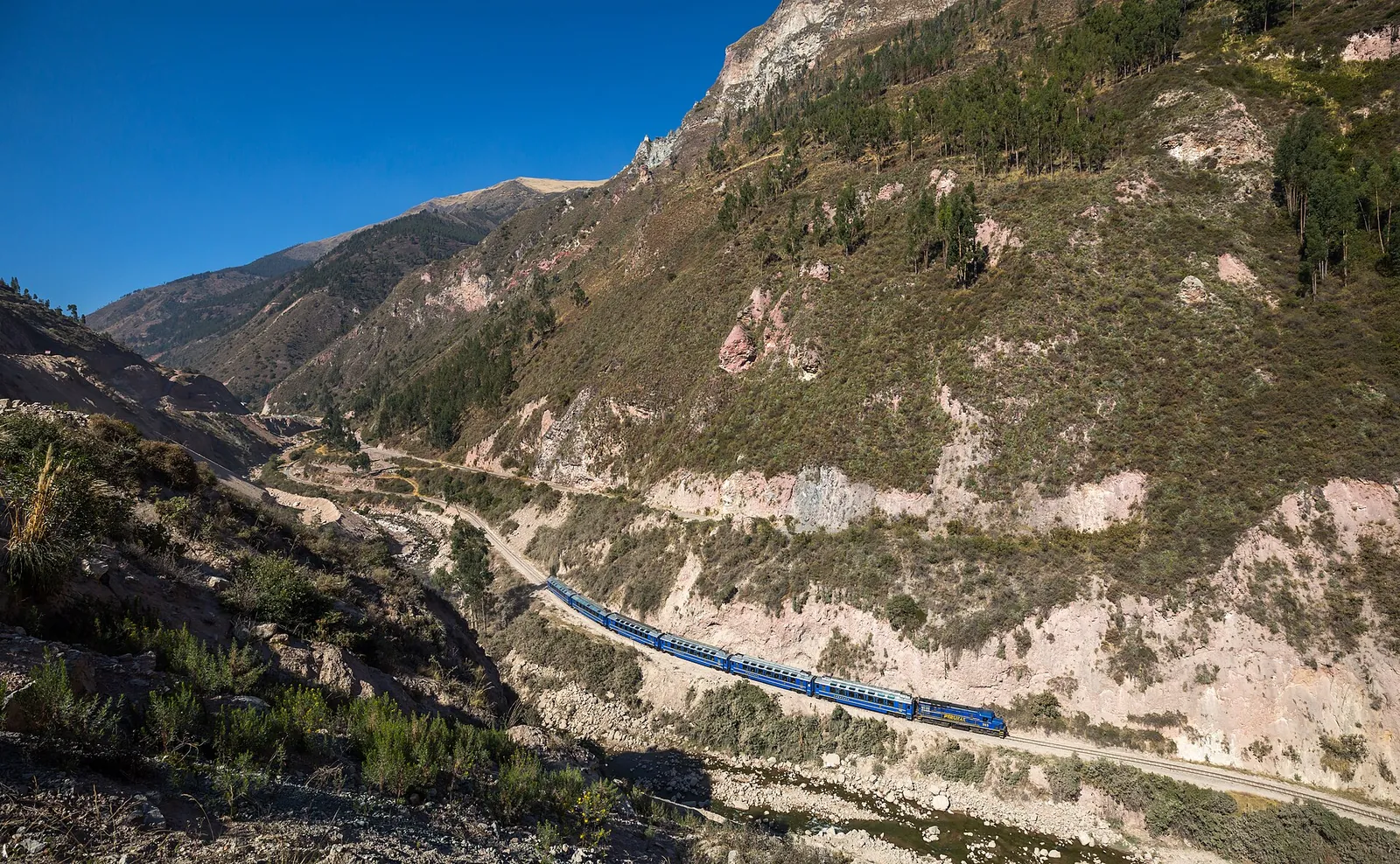 A blue PeruRail train winding through the valley