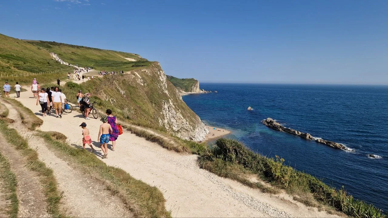 A chalk footpath running along green cliffs above the blue sea on the Jurassic Coast, with walkers heading down