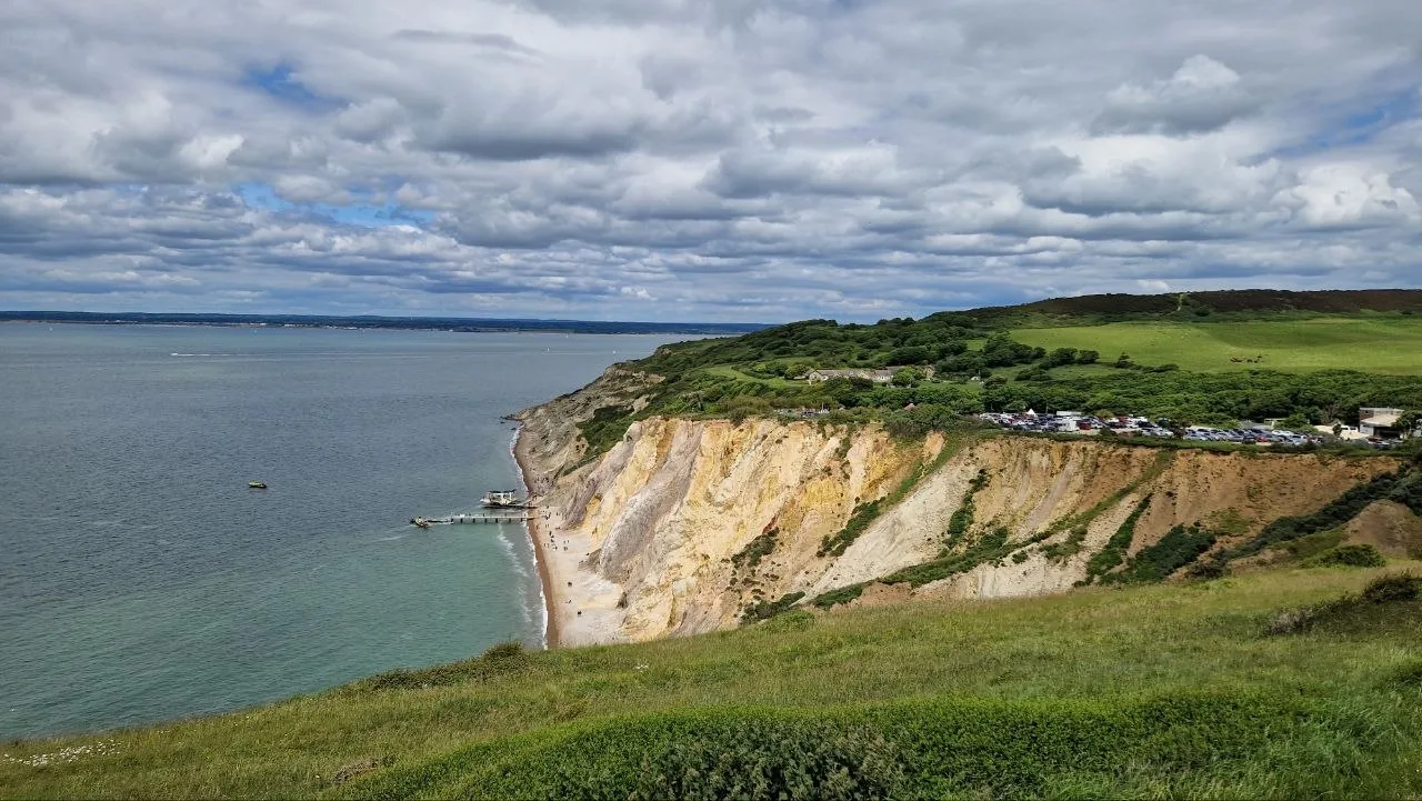 White and ochre sea cliffs above a narrow beach and a small pier on the Isle of Wight, with green clifftop fields