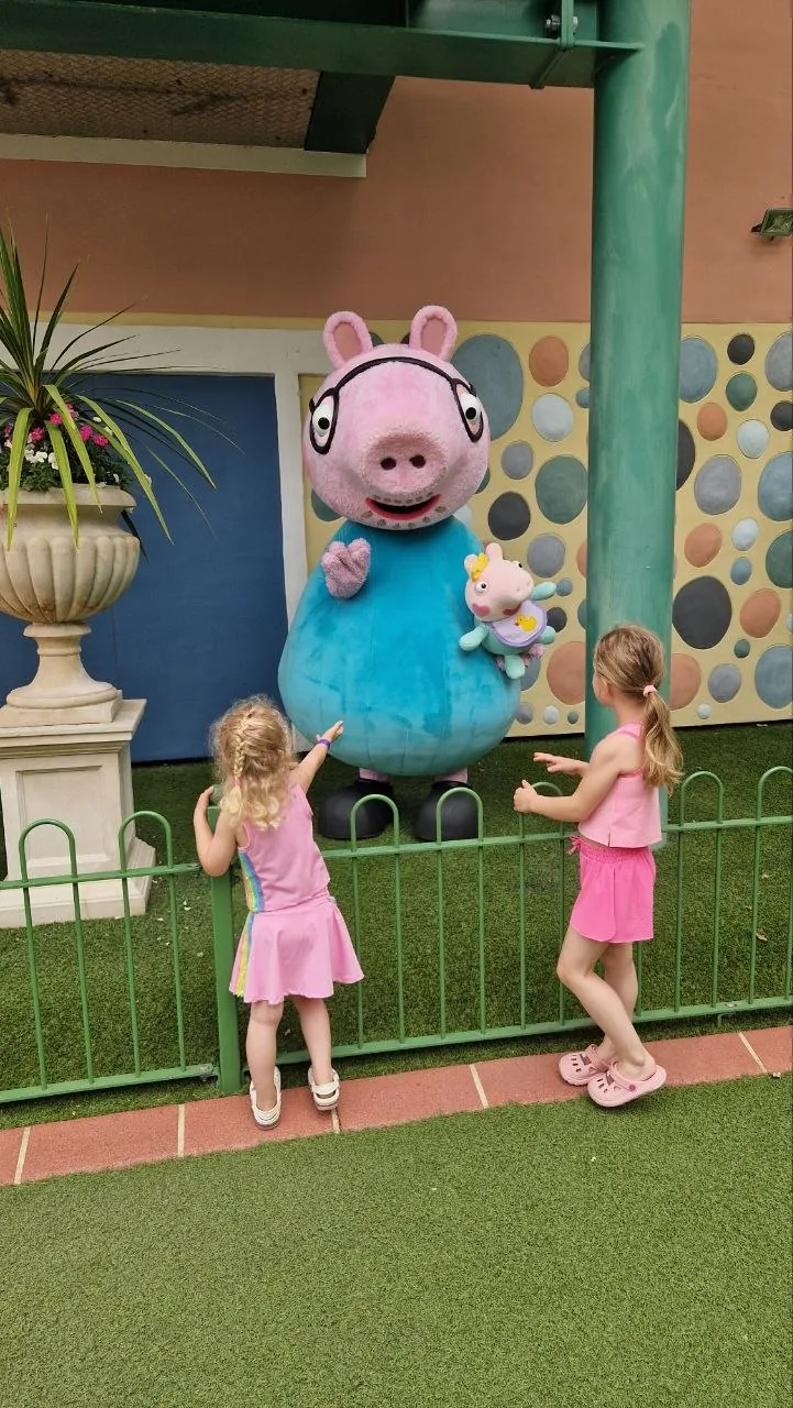 Two little girls in pink waving at George Pig outside a house in Peppa Pig World